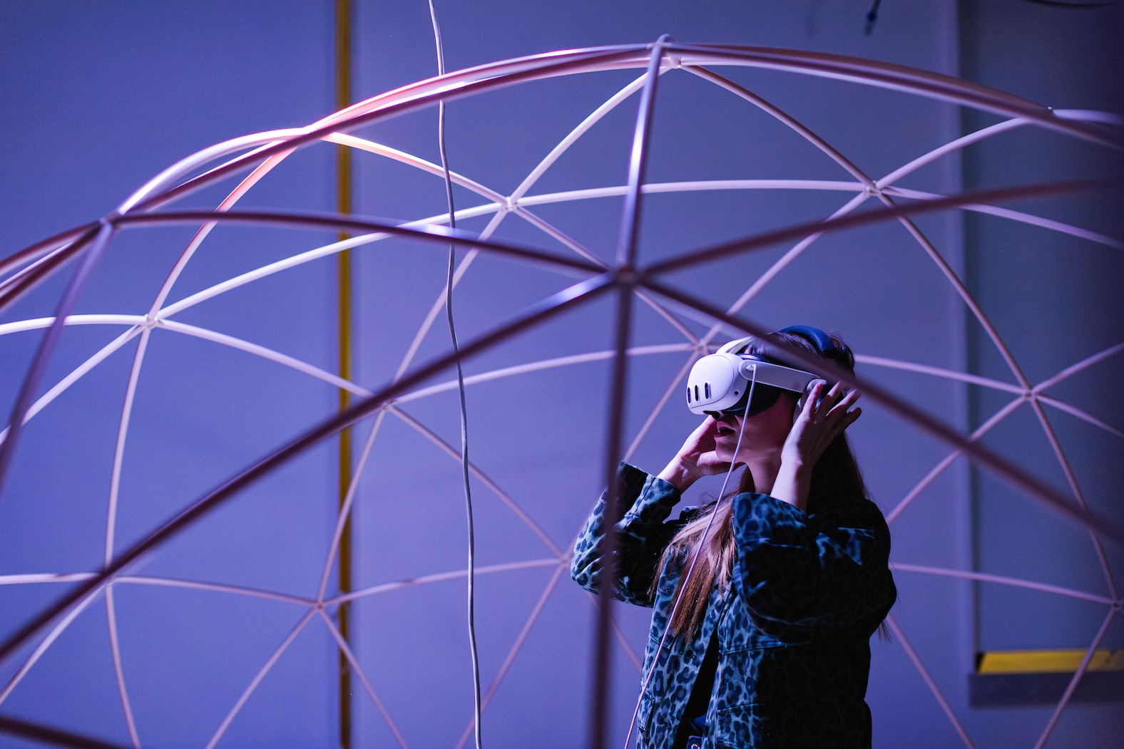 Επισκέπτρια του Athens Digital Arts Festival φοράει γυαλιά εικονικής πραγματικότητας (VR), εξερευνώντας μια διαδραστική εγκατάσταση με φουτουριστικό φωτισμό και γεωδαιτική δομή.